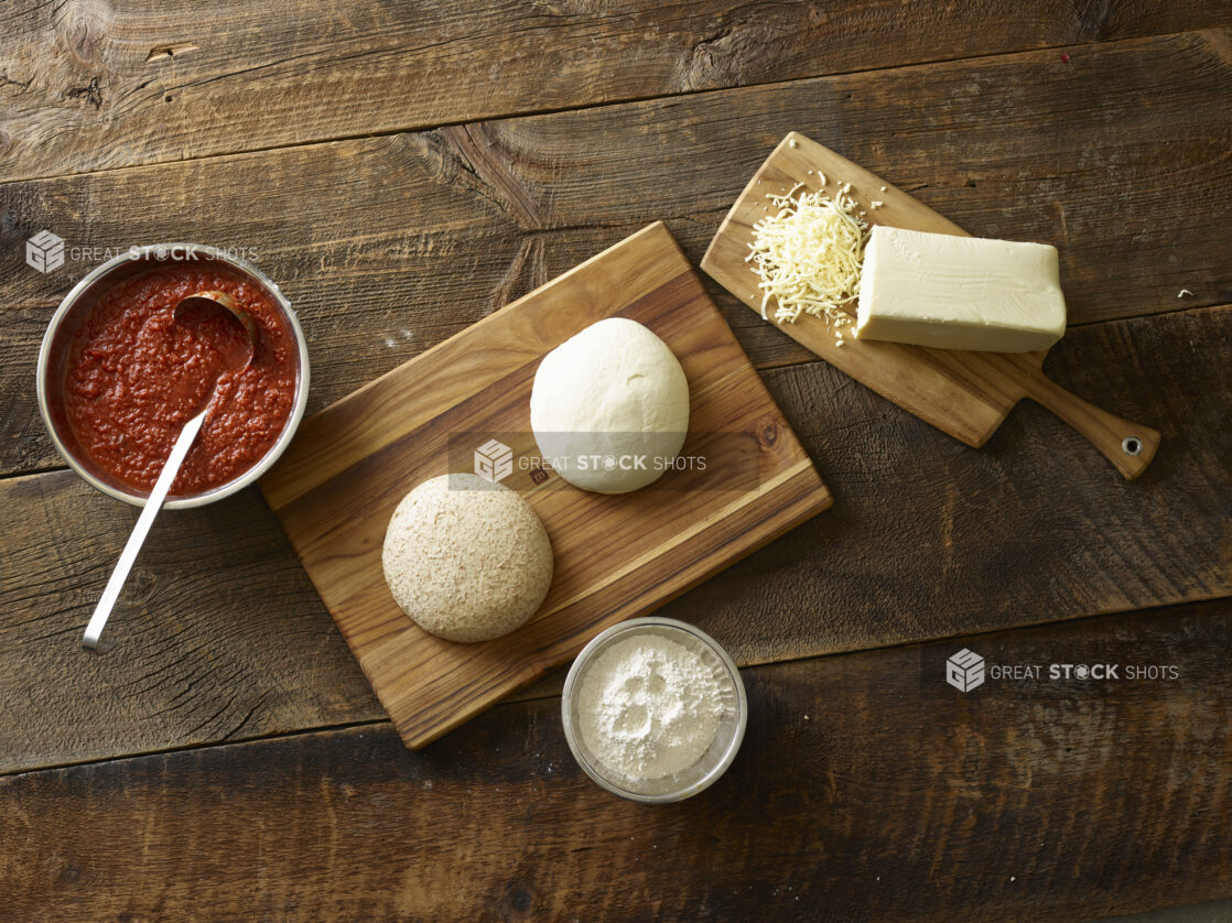 Pizza dough on a wooden cutting board, pizza sauce in a bowl and cheese brick, bowl of flour on a aged wooden table with an overhead view