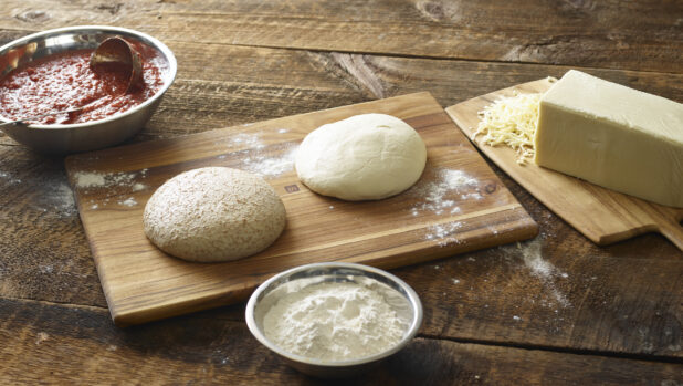 Pizza dough on a wooden cutting board with a bowl of sauce, cheese and a bowl of flour on aged wooden table