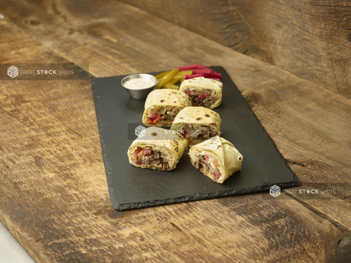 Shawarma bites on a slate platter with pickles, pickled turnip & dip on a wooden background slightly angled