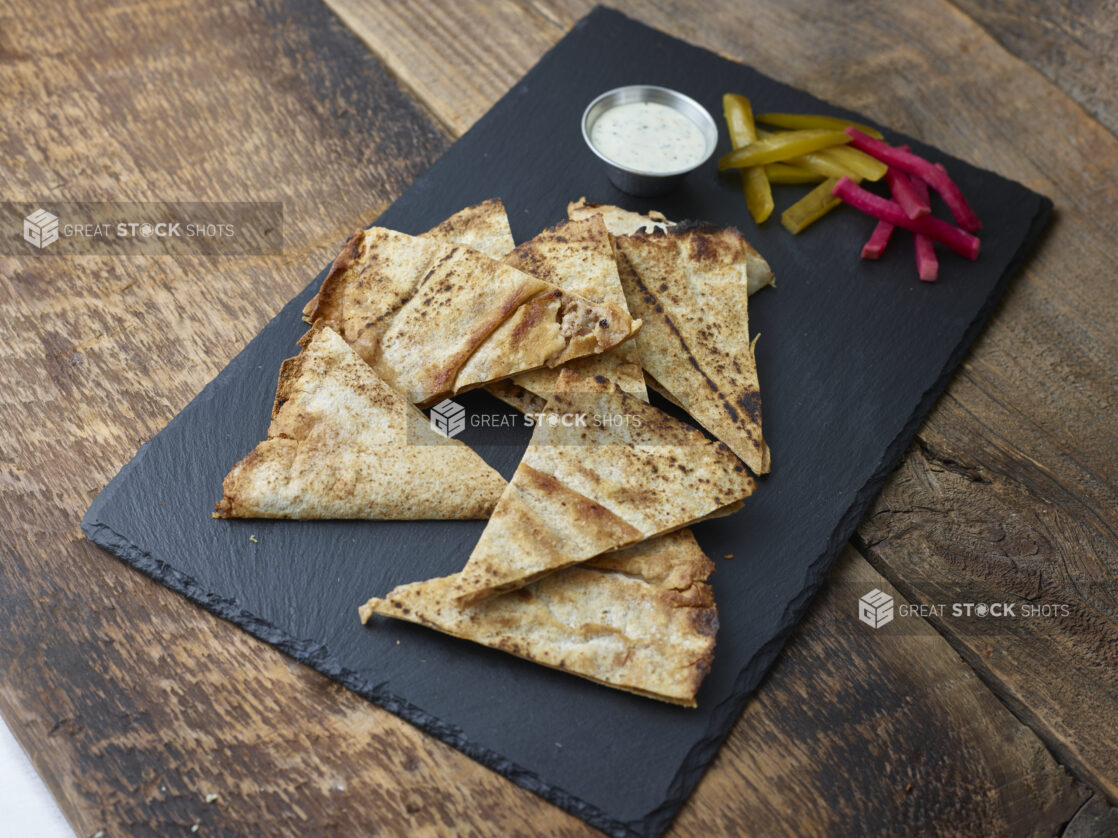 Lahmajeen/middle eastern pizza on a slate platter with pickles, turnip and dip on a wooden table on a 45 degree angle