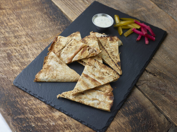 Lahmajeen/middle eastern pizza on a slate platter with pickles, turnip and dip on a wooden table on a 45 degree angle