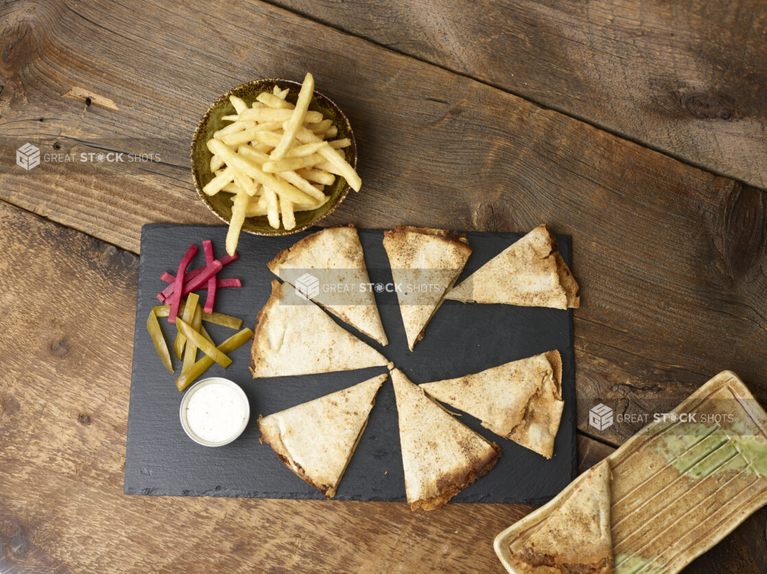 Overhead view of a lahmajeen/middle eastern pizza with french fries, pickles, turnip and dip on a slate platter on a wooden background