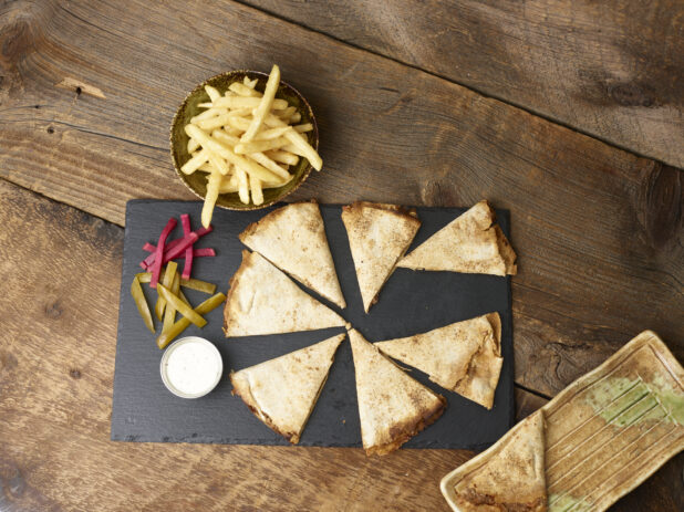 Overhead view of a lahmajeen/middle eastern pizza with french fries, pickles, turnip and dip on a slate platter on a wooden background