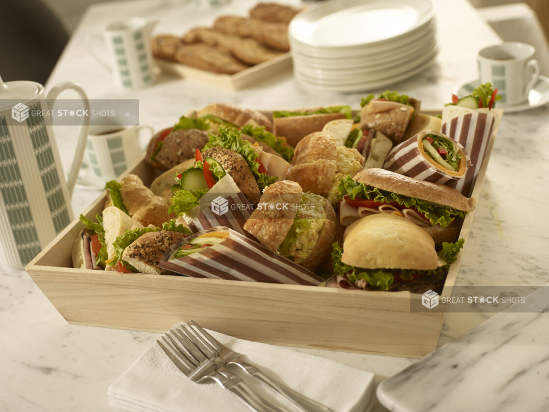 Assorted sandwiches and wraps in a wood catering box with side plates, coffee, forks, cookie tray, coffee cups on a white marble background