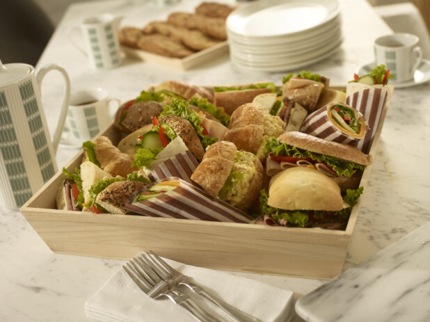 Assorted sandwiches and wraps in a wood catering box with side plates, coffee, forks, cookie tray, coffee cups on a white marble background