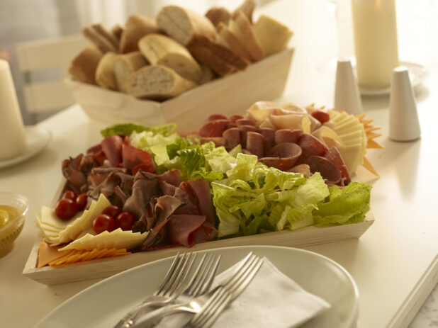 Assorted deli meat on a wood catering tray with assorted cheeses, lettuce and cherry tomatoes with assorted breads and rolls in a wood catering box on a white table with table accessories