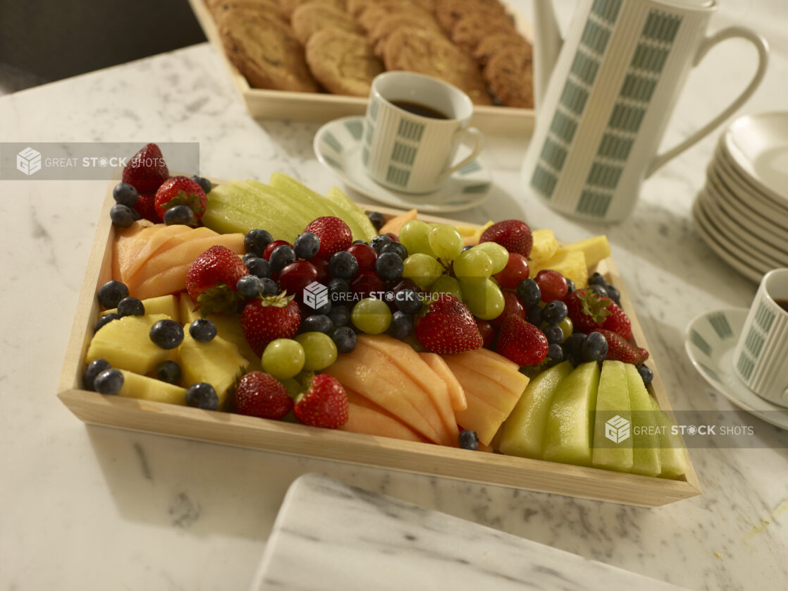 Fresh fruit tray with sliced honeydew melon, cantaloupe, pineapple, grapes, strawberries and blueberries on a wood catering tray with assorted cookies on a wood catering tray with a coffee carafe, coffee cups and white side plates on a marble background