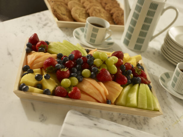 Fresh fruit tray with sliced honeydew melon, cantaloupe, pineapple, grapes, strawberries and blueberries on a wood catering tray with assorted cookies on a wood catering tray with a coffee carafe, coffee cups and white side plates on a marble background
