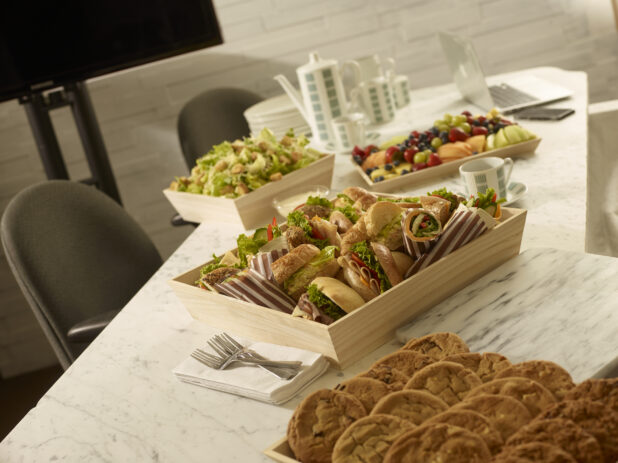 Assorted platters, deli sandwich platter, fresh fruit tray, caesar salad and assorted cookie tray, all on wood catering trays/boxes in a office meeting setting
