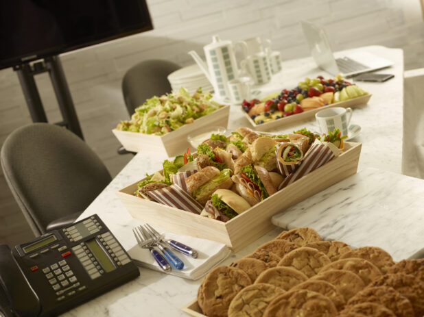 Various platters, assorted deli sandwiches, caesar salad, assorted fresh fruit and assorted cookies all on wood catering trays on a marble table in a office meeting setting