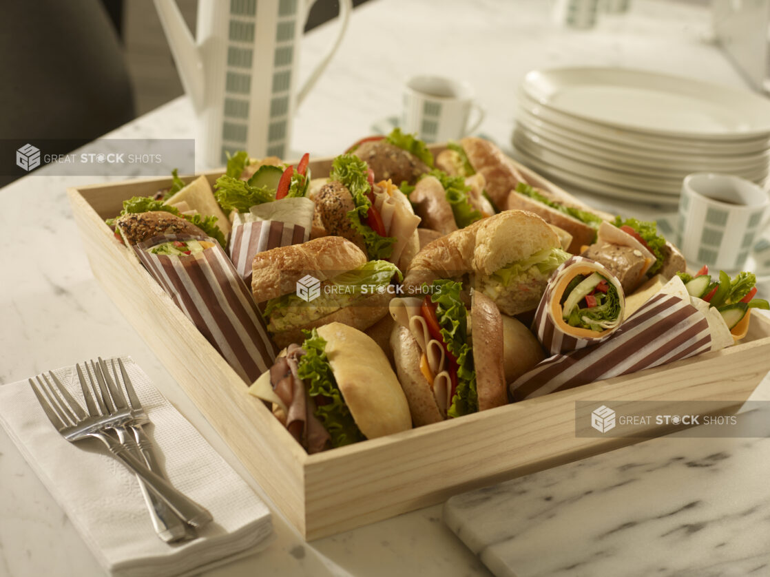 Assorted deli sandwiches in a wood catering box with coffee carafe, coffee cups, side plates, forks and napkins on a marble background