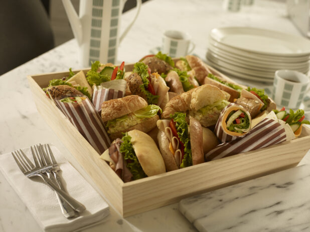 Assorted deli sandwiches in a wood catering box with coffee carafe, coffee cups, side plates, forks and napkins on a marble background