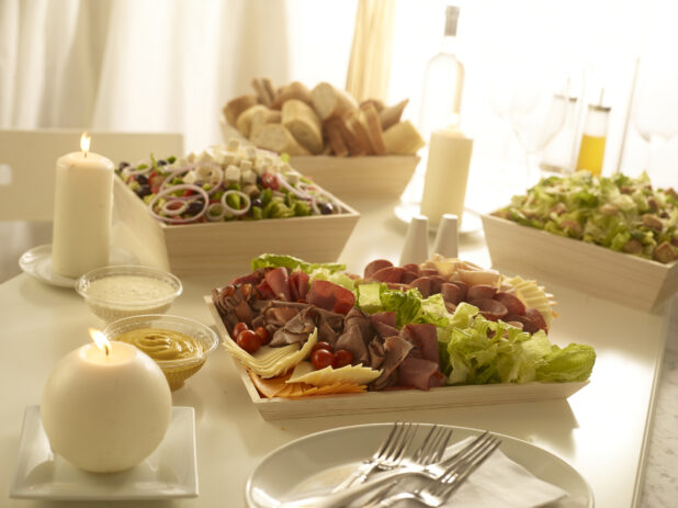 Deli meat tray, Greek salad, Caesar salad, bread and rolls all in wood catering trays and boxes in an elegant table setting