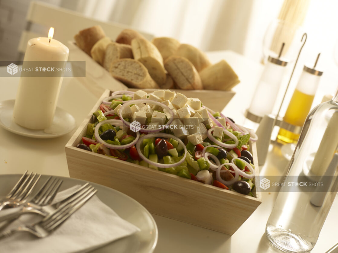 Wood catering tray will with Greek salad with a tray of sliced buns in the background on a white countertop in a home