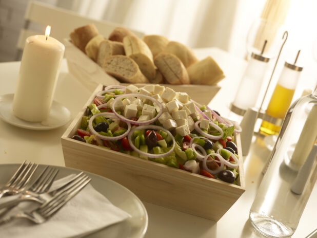 Wood catering tray will with Greek salad with a tray of sliced buns in the background on a white countertop in a home