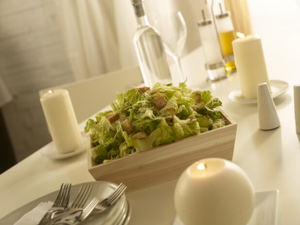 Caesar salad in a wood catering box in a elegant table setting with candles and other accessories