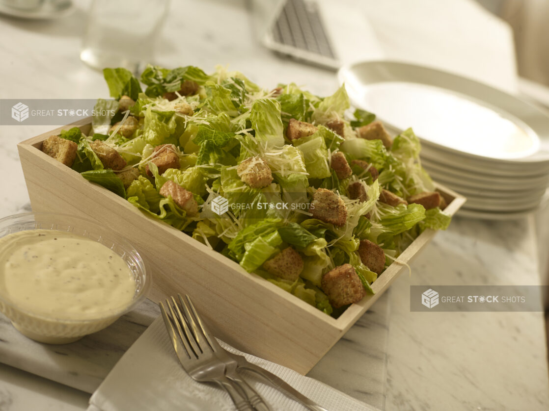 Caesar salad with croutons, parmesan cheese in a wood catering box and dressing on the side with side plates and forks on a white marble background