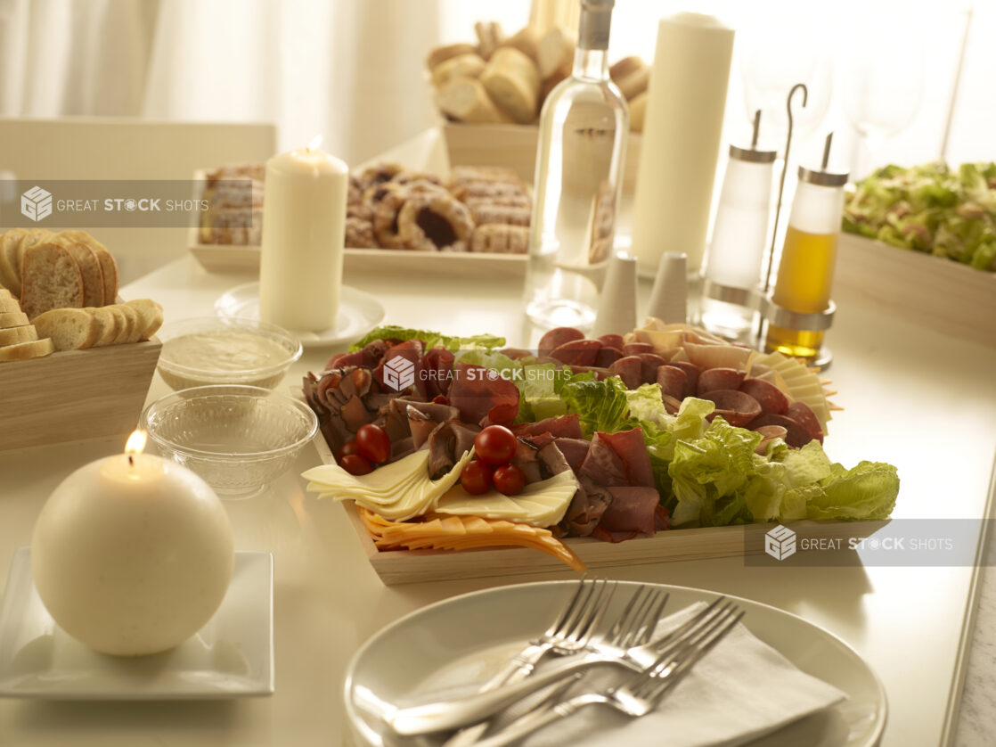 Assorted deli meat platter with lettuce, sliced cheese and grape tomatoes, assorted danishes, caesar salad and assorted breads and rolls in an elegant table setting