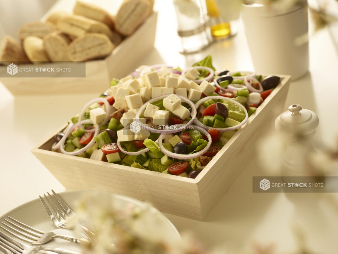 Greek salad with red onion, black olives, cucumber, tomatoes in a wood catering box with a wood box of bread, side plates and forks in the background and foreground