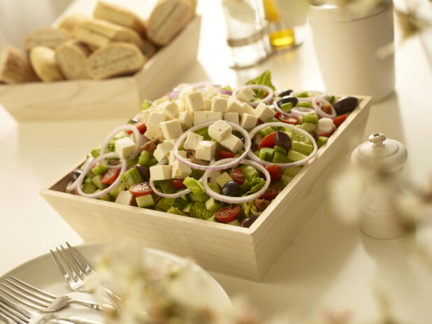 Greek salad with red onion, black olives, cucumber, tomatoes in a wood catering box with a wood box of bread, side plates and forks in the background and foreground