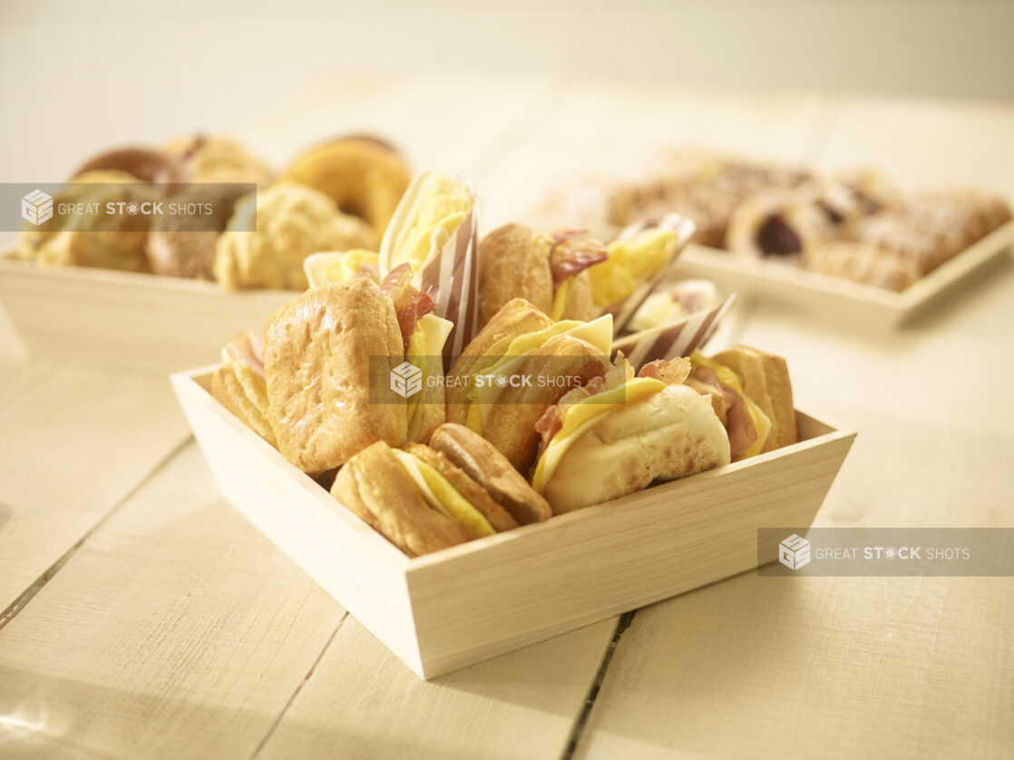 Assorted breakfast sandwiches in a wood catering box with assorted pastries in and on wood catering trays/boxes in the background on a wooden background