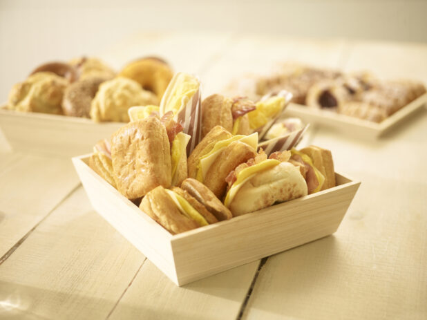 Assorted breakfast sandwiches in a wood catering box with assorted pastries in and on wood catering trays/boxes in the background on a wooden background