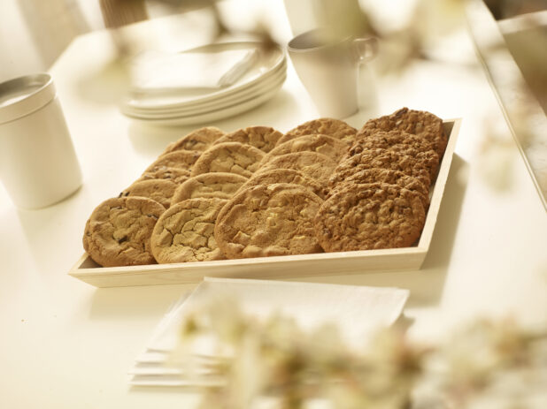 Assorted cookies on a wood catering tray with side plates, mugs and napkins on a white table