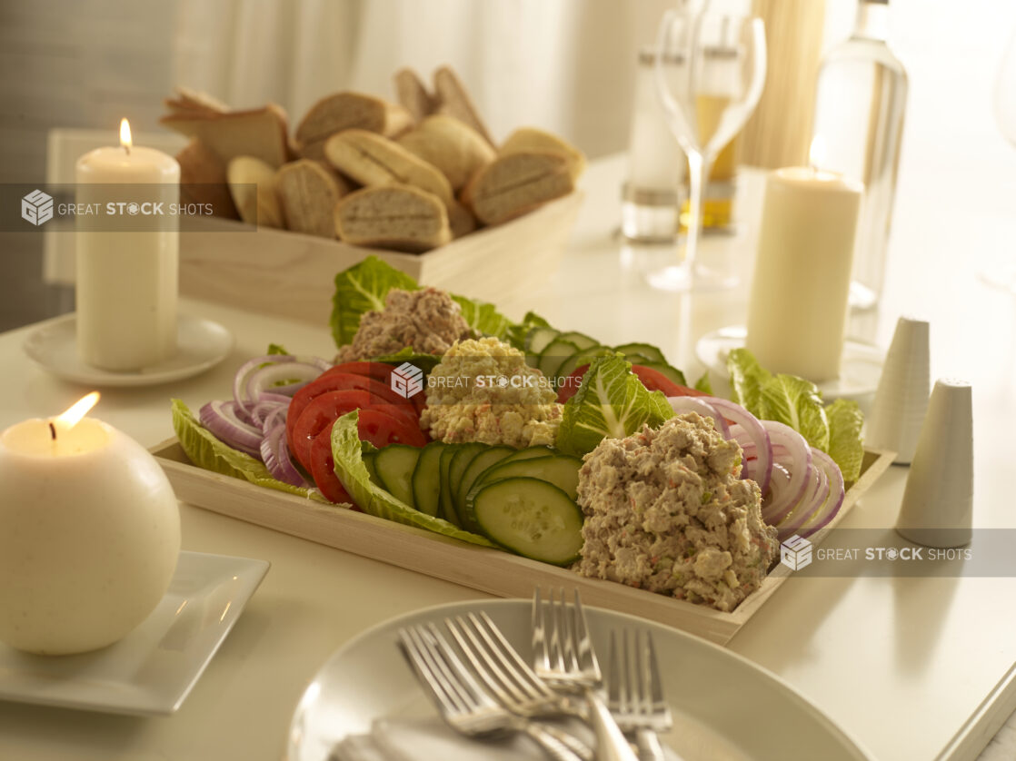 Dairy platter with egg salad, tuna salad and salmon salad in mounds with sliced red onions, cucumbers and tomatoes on a wood catering tray with a wood catering box of assorted breads and rolls, in an elegant table setting