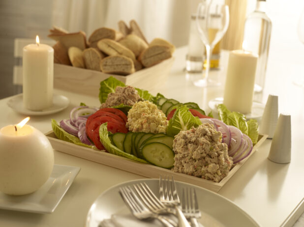 Dairy platter with egg salad, tuna salad and salmon salad in mounds with sliced red onions, cucumbers and tomatoes on a wood catering tray with a wood catering box of assorted breads and rolls, in an elegant table setting
