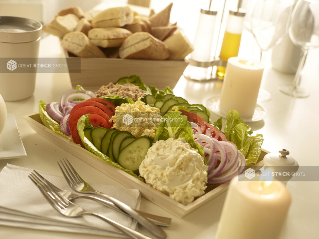 Egg salad, tuna salad and cream cheese in mounds with sliced cucumber, sliced tomatoes and sliced red onions in a wood catering box with a wood box of bread in the background with table accessories