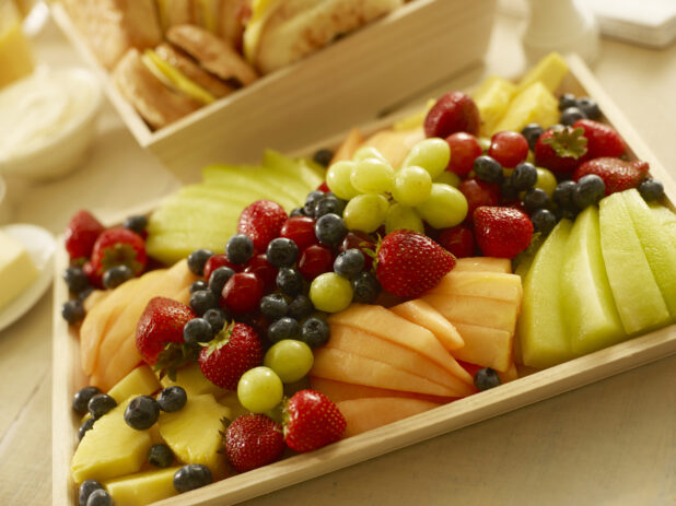 Fresh fruit on a wood catering tray with sliced melon, cantaloupe, pineapple, grapes, strawberries and blueberries with other breakfast items in the background
