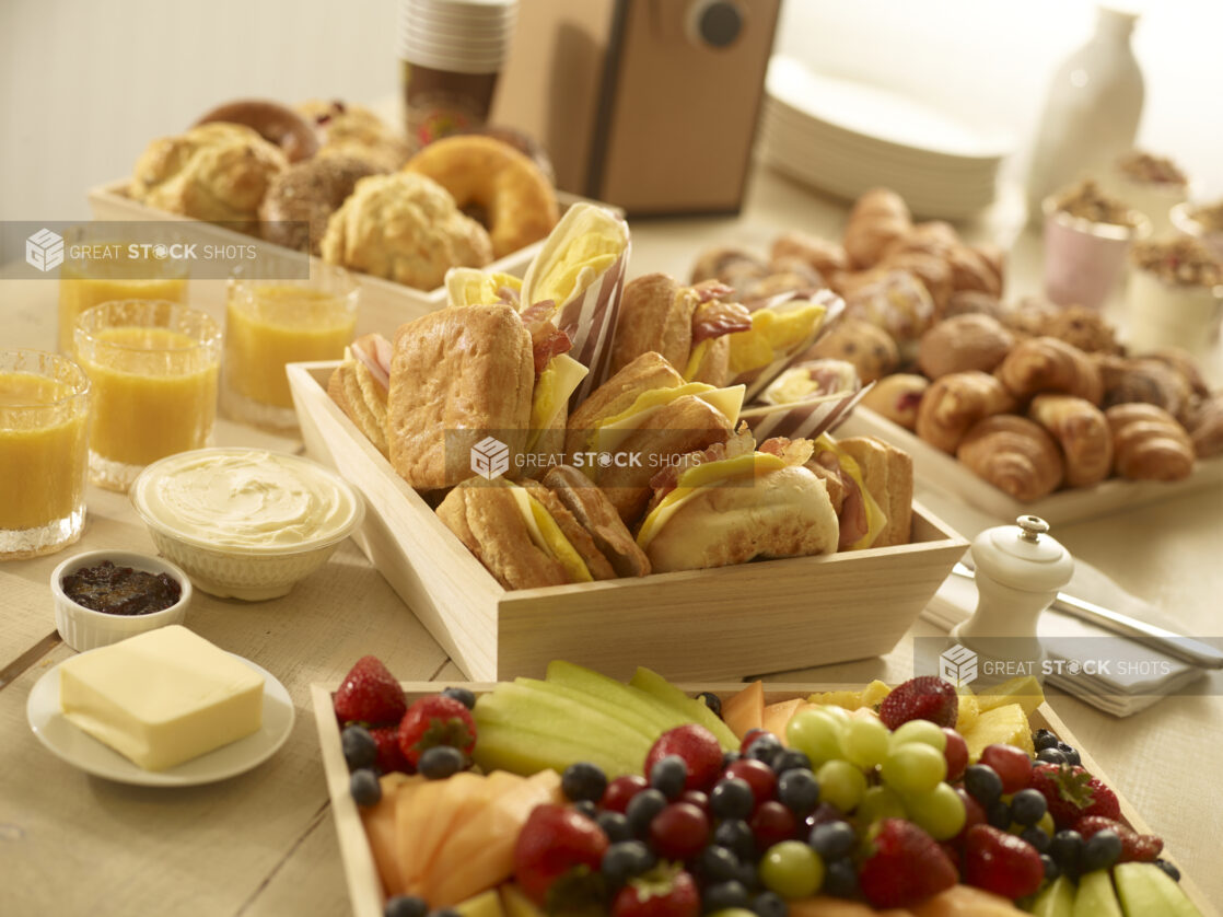 Breakfast sandwiches, fresh fruit tray, assorted pastries, scones and bagels with yogurt parfaits, jam, cream cheese, butter and glasses of orange juice surrounding