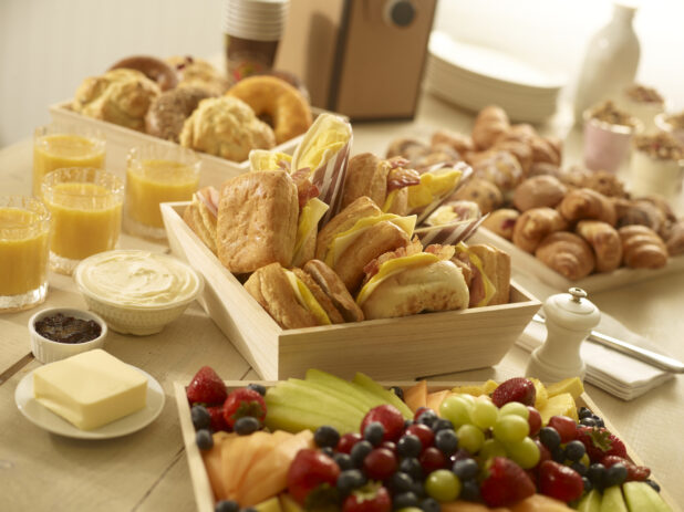 Breakfast sandwiches, fresh fruit tray, assorted pastries, scones and bagels with yogurt parfaits, jam, cream cheese, butter and glasses of orange juice surrounding