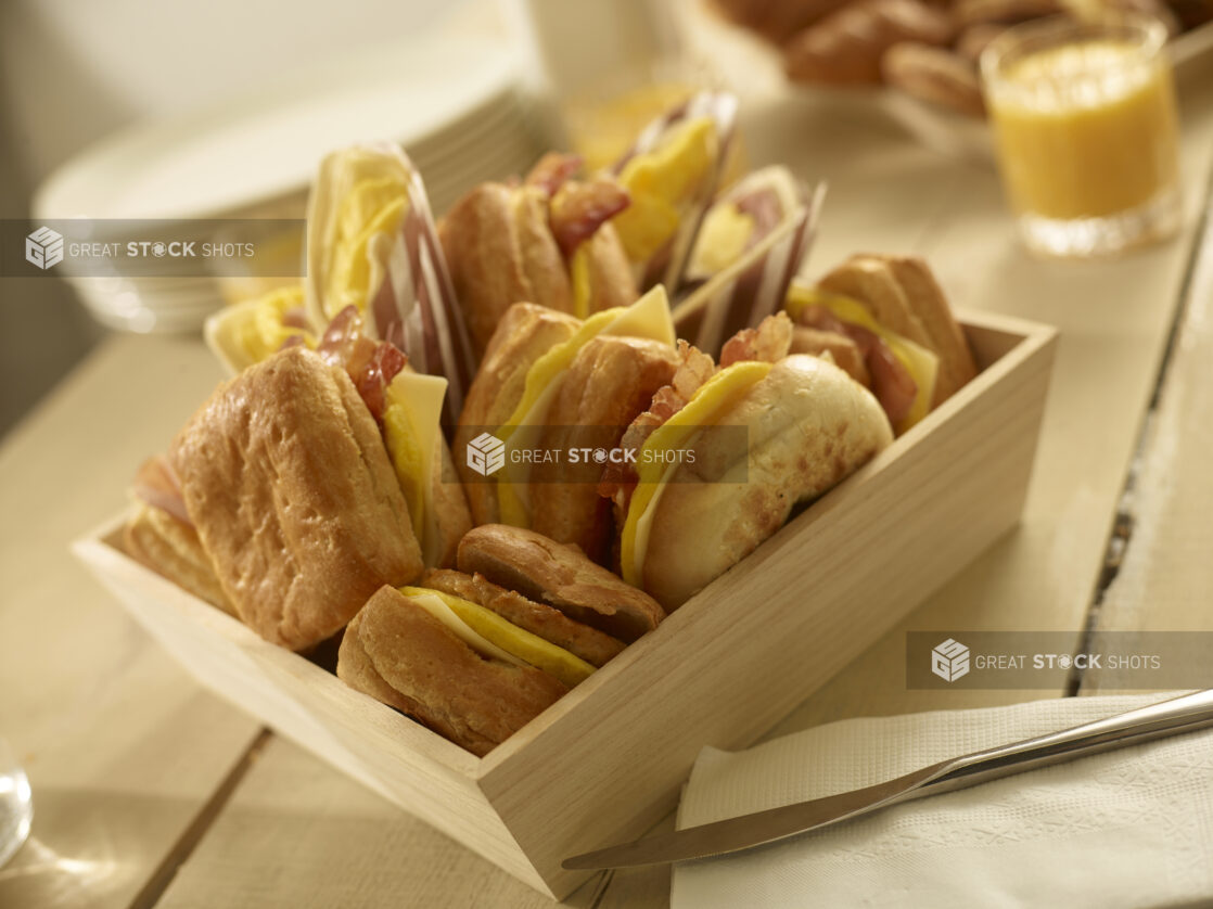 Close up view of assorted breakfast sandwiches in a wood catering box with additional breakfast items in the background, close-up