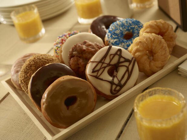 Assorted donuts on a rectangular wood catering tray, with glasses of orange juice in the foreground and background