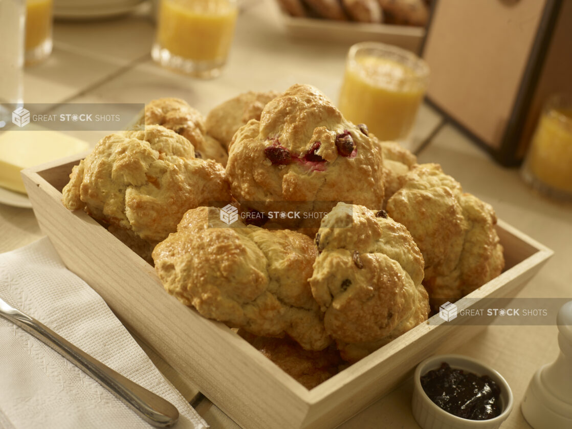 Close up view of assorted breakfast scones/tea biscuits in a wood catering box with a ramekin of jar, butter and small glasses of orange juice in the background