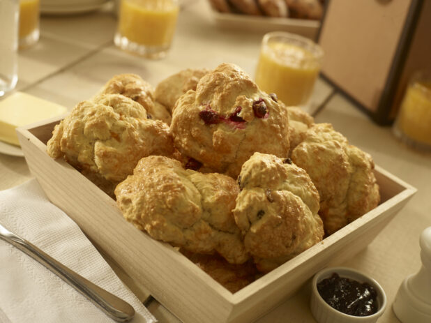 Close up view of assorted breakfast scones/tea biscuits in a wood catering box with a ramekin of jar, butter and small glasses of orange juice in the background
