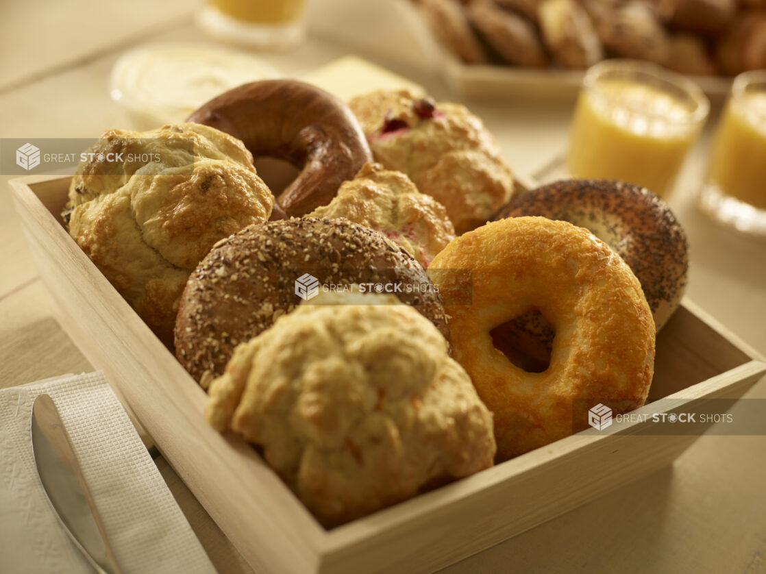 Close up view of assorted bagels and tea biscuits/scones in a wood catering box with other breakfast items in the background