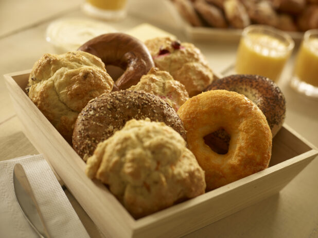 Close up view of assorted bagels and tea biscuits/scones in a wood catering box with other breakfast items in the background