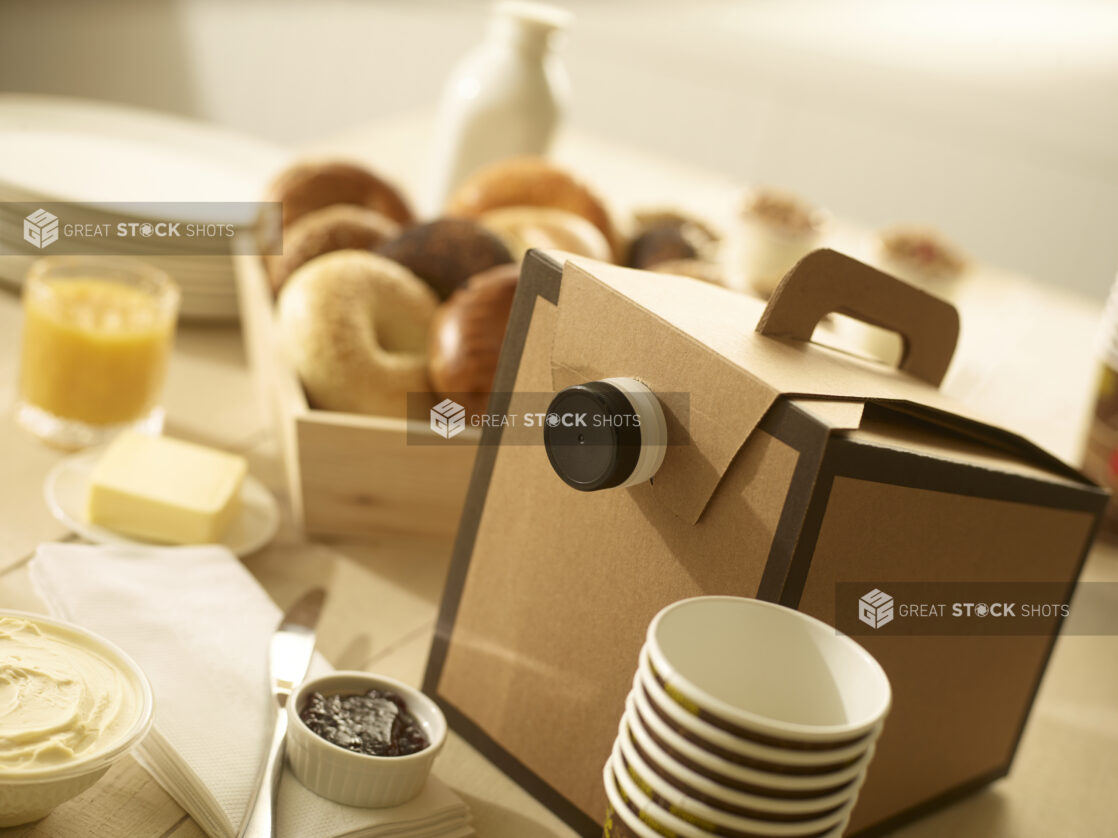 Disposable coffee cups and disposable coffee dispenser with assorted whole, uncut bagels in a wood catering box with butter, jam, cream cheese, on the side