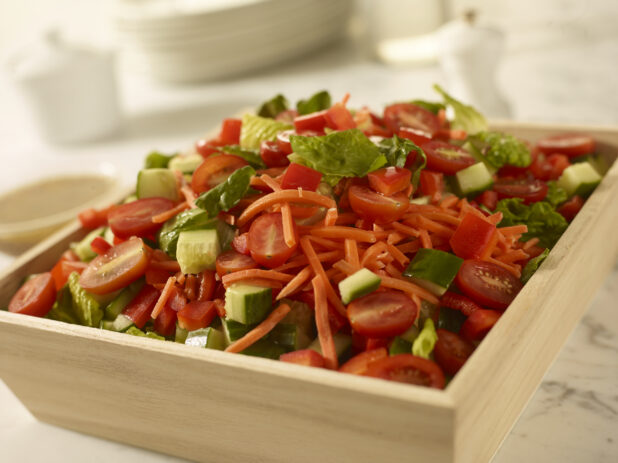 Garden salad with cucumber, tomato and shredded carrots in a wood catering box, close up view on a white a marble background