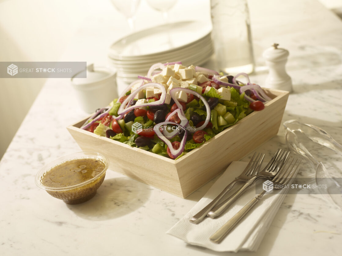 Greek salad in a wood catering box with dressing, cutlery, side plates and napkins on the side