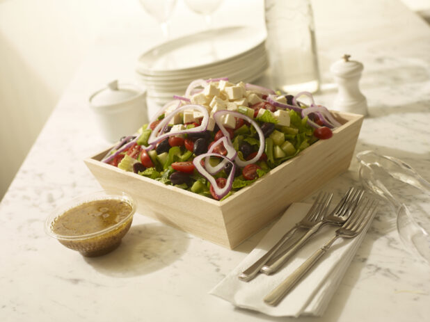 Greek salad in a wood catering box with dressing, cutlery, side plates and napkins on the side