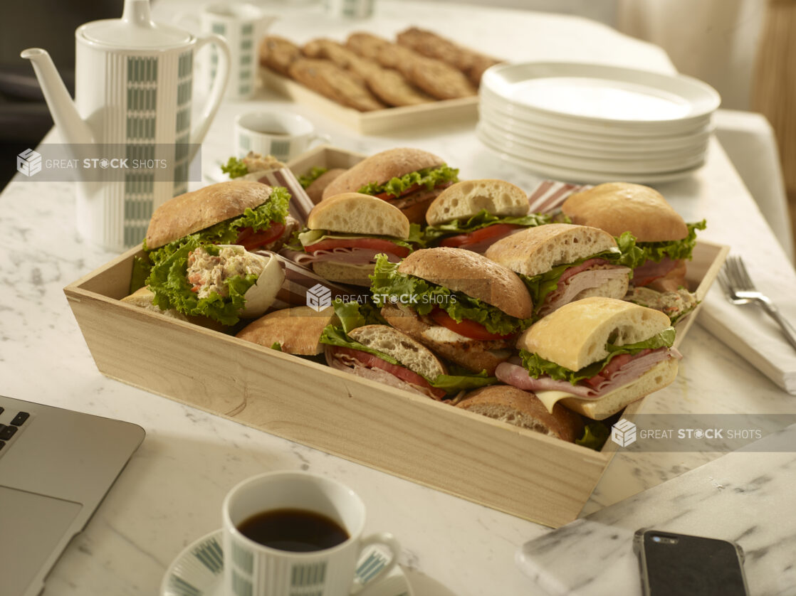 Assorted deli sandwiches in a wood catering box with coffee carafe, coffee cups, side plates, forks and napkins on a marble background in a meeting settng