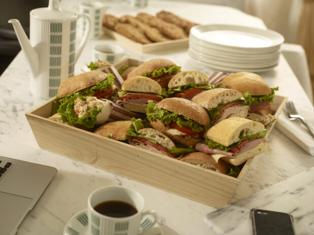 Assorted deli sandwiches in a wood catering box with coffee carafe, coffee cups, side plates, forks and napkins on a marble background in a meeting settng