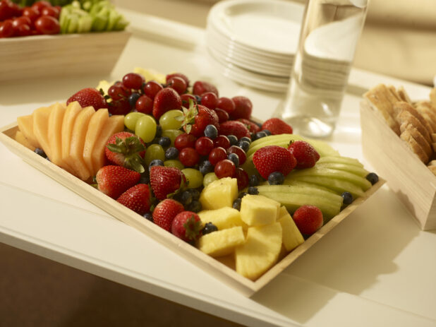 Fresh fruit platter with sliced melon, cantaloupe, pineapple, strawberries, grapes and blueberries on a rectangular wood catering tray with side plates, bread box and veggies and dip in the background