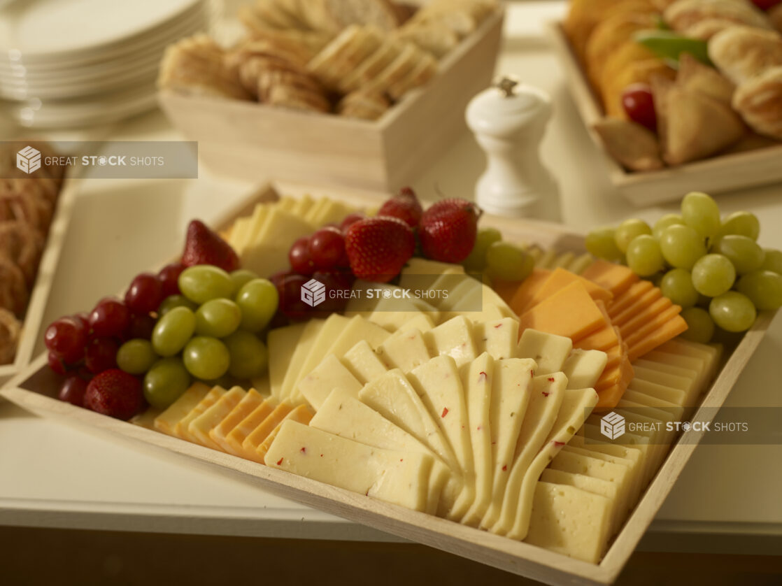 Cheese platter on a wood catering tray with assorted cheeses, grapes and strawberries with other party platters surrounding