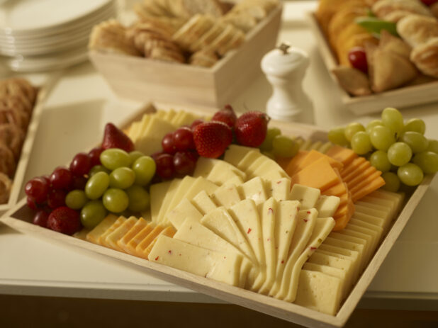 Cheese platter on a wood catering tray with assorted cheeses, grapes and strawberries with other party platters surrounding