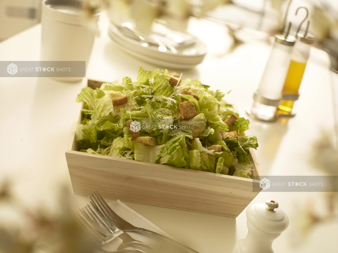 Caesar salad with croutons and parmesan cheese in square wood catering box with table accessories in the foreground and background