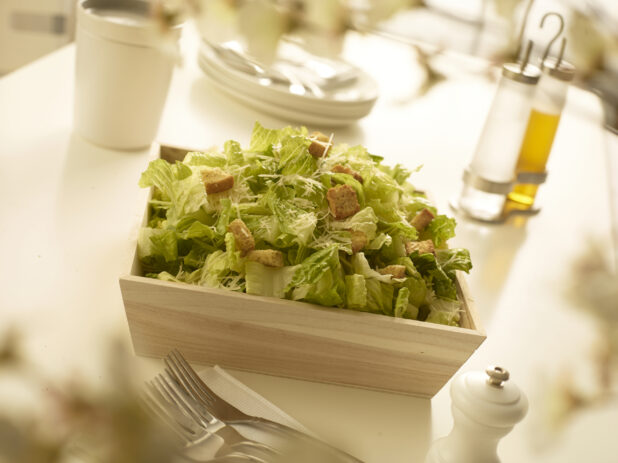 Caesar salad with croutons and parmesan cheese in square wood catering box with table accessories in the foreground and background